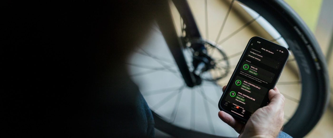 SRAM AXSアプリが表示されたスマートフォンを手に持ち、自転車のRival電子式ドライブトレインの設定を行っている人物。.