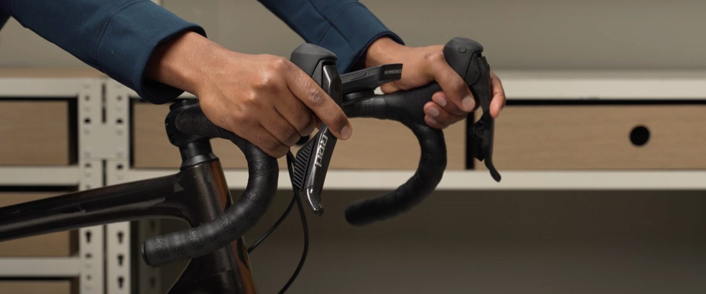 変速レバーとサイクルコンピュータが取り付けられた自転車のドロップハンドルを、両手で握っている様子。.