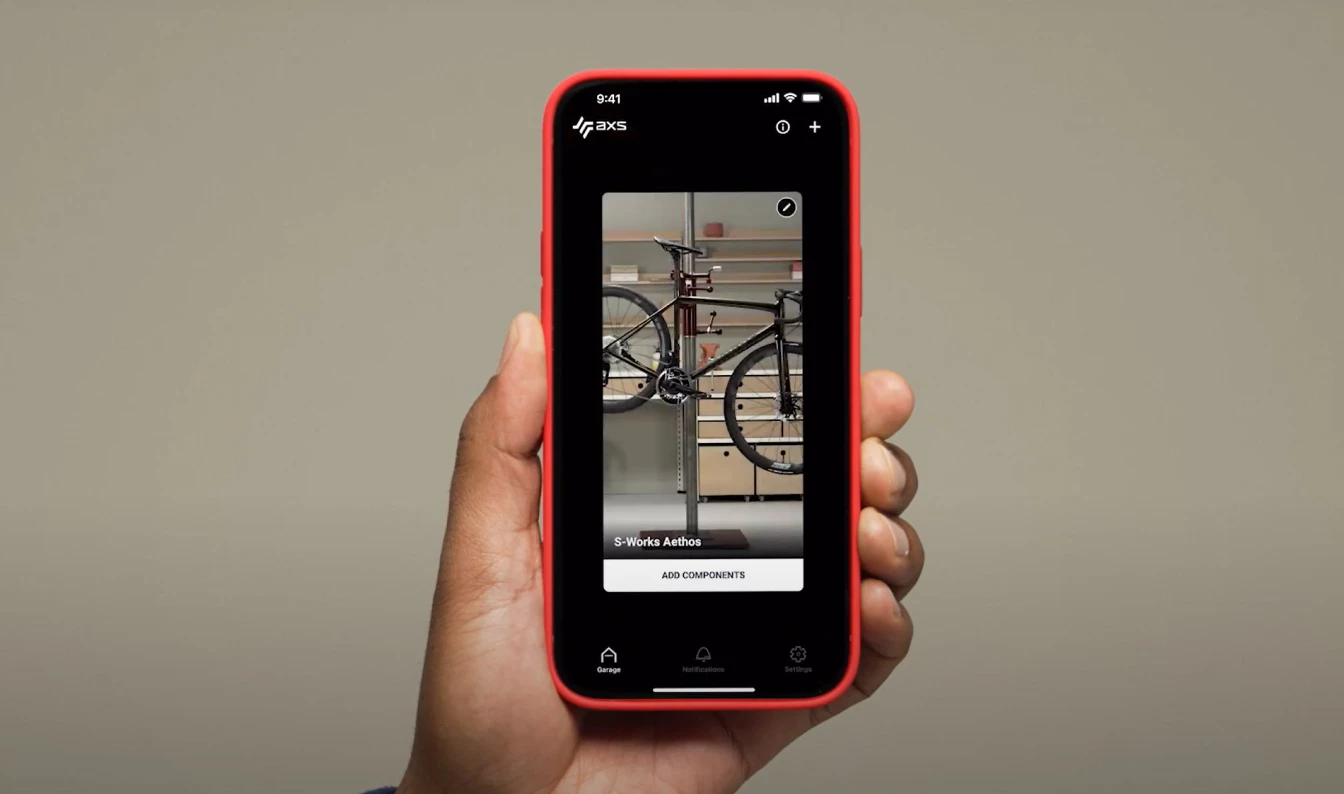 SRAM AXSアプリが表示されたスマートフォンを手に持った人物が、修理スタンドに載せられたS-Works Aethosの自転車と、コンポーネントを追加ボタンを映し出している。.