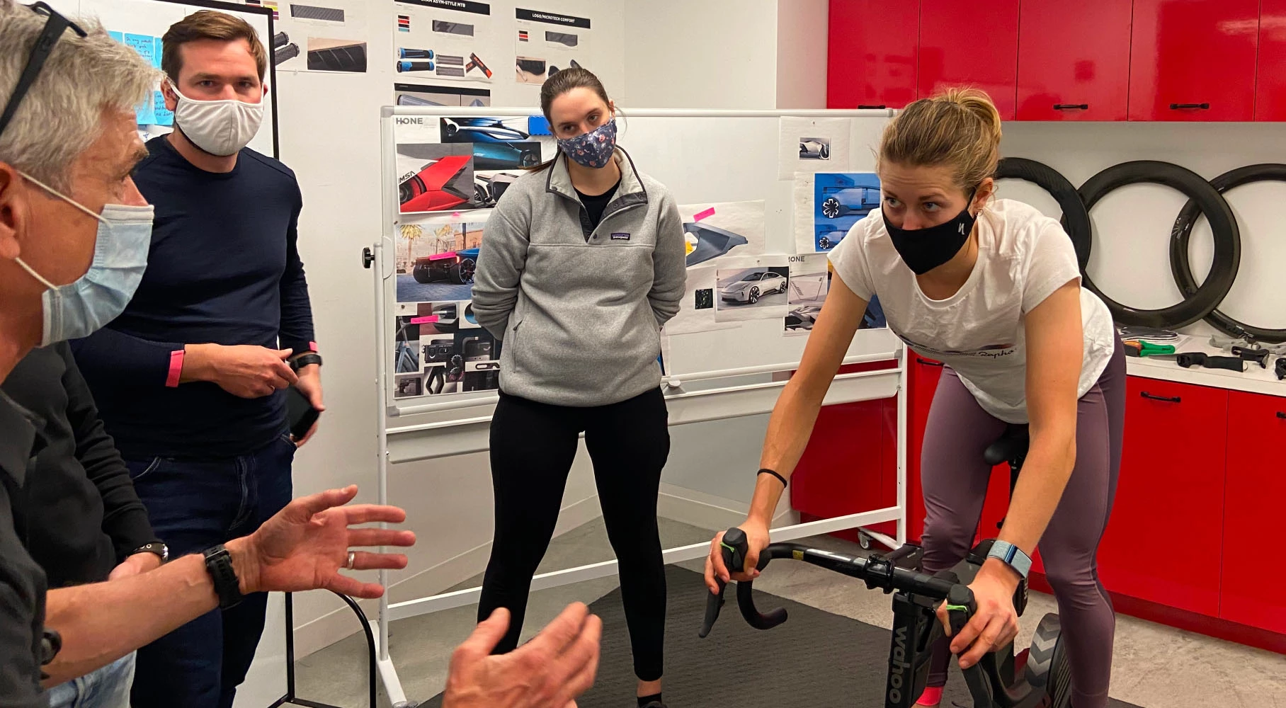 スポーツウェア姿の女性がエアロバイクをこいでいる。その様子を、ホワイトボードにデザインスケッチが書かれたワークショップの会場で、3人が見守っている。.