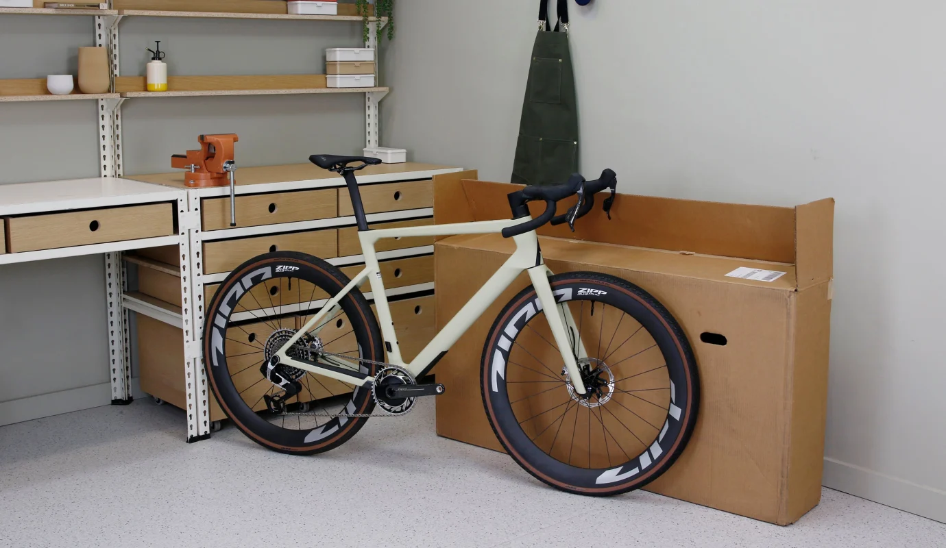 Zippのホイールを装着した明るい色のロードバイクが、作業場の配送用箱の隣に停められている。.
