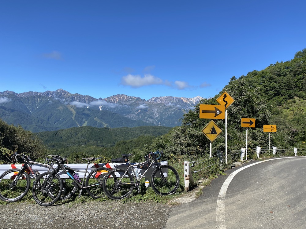 曲がりくねった道路標識がいくつもある山道の脇に3台の自転車が停まっている。背景には澄み切った青空の下、森に覆われた丘と山が見える。.