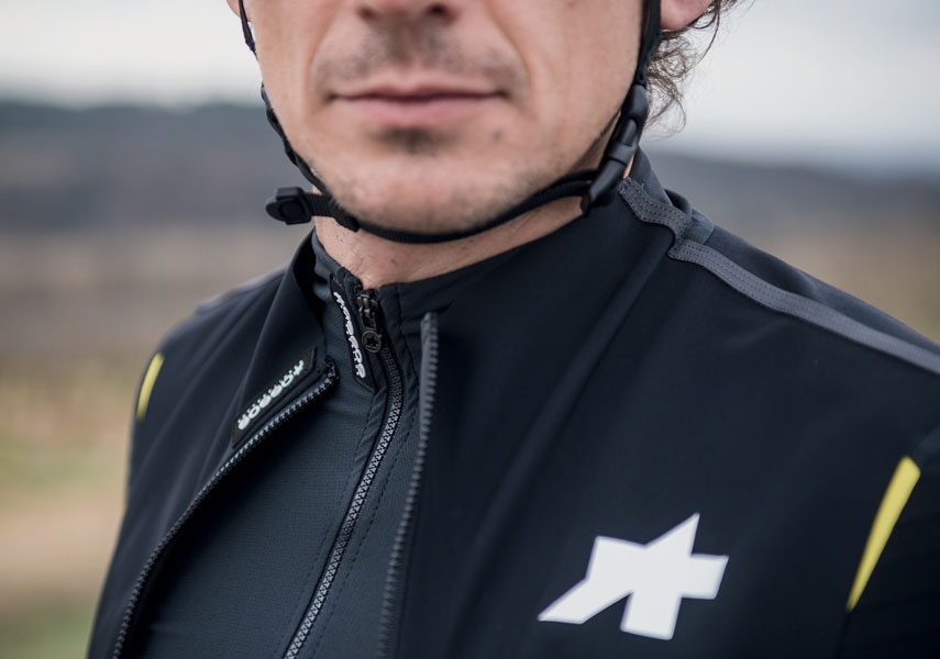 白い幾何学模様のロゴが入った黒のサイクリングジャケットを着用し、ヘルメットのあご紐を締めた人物のクローズアップ、屋外。.