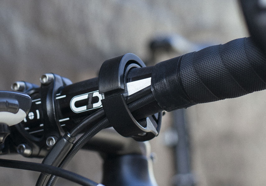 自転車のハンドルバーのクローズアップ。黒いテープ巻き、ギアケーブル、ステム付近に取り付けられた円形のプラスチック製アクセサリーが見える。.