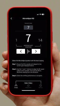 ある手が、サイクリングアプリのMicroAdjust RD画面が表示されたスマートフォンを持ち、ギア選択の操作ボタンと、リアディレイラーの調整手順を3つのステップで示した画面が表示されている。.