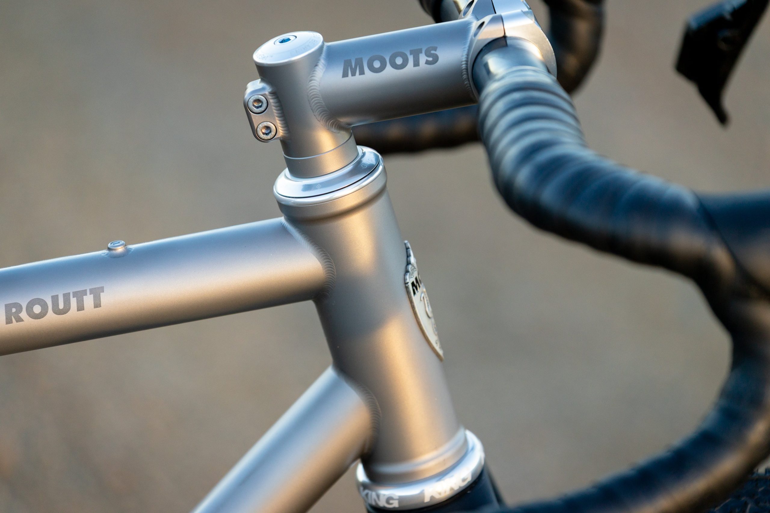 A33A0256 ROUTT製の金属製自転車フレームのクローズアップ写真。ヘッドチューブ、ステム、そしてグリップが巻かれたハンドルバーが見える。.