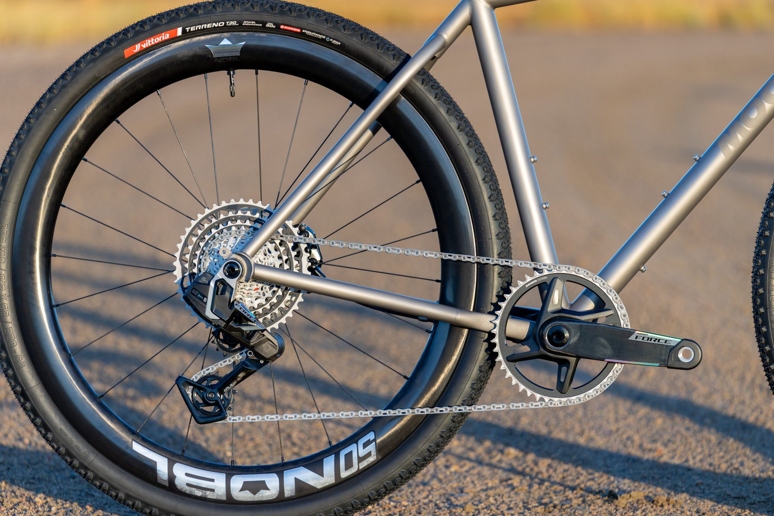 A33A0271 ROUTT製のチタン製グラベルバイクフレームのクローズアップ。シングルチェーンリングのドライブトレイン、リアディレイラー、そしてノッビータイヤを装着したディープセクションのカーボンホイールが特徴だ。.