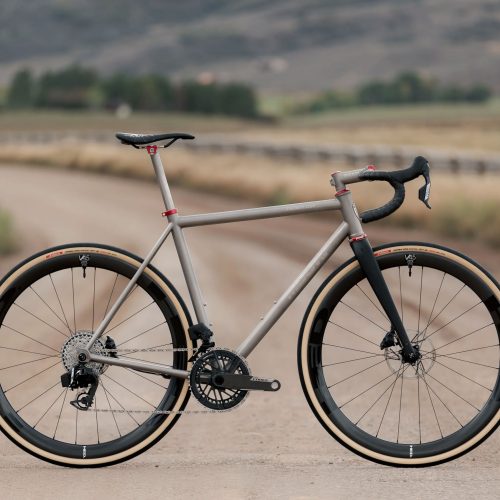 チタンフレーム、ブラックカーボンフォーク、ディープセクションホイール、ドロップハンドルを備えたCOUNTY LINE ALL-ROAD BIKEが未舗装路を走っている。.