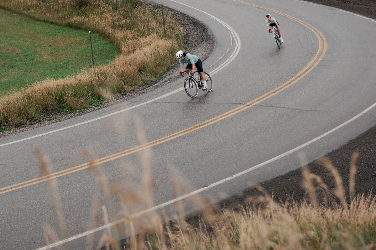 2人のサイクリストが、アスファルト道路のカーブをCOUNTY LINE ALL-ROAD BIKESに乗って走っている。.