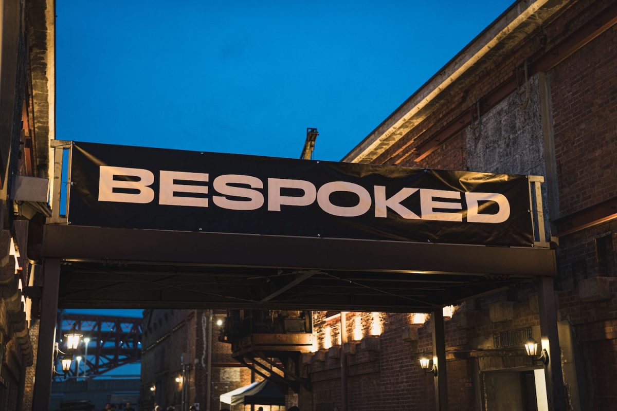 2つのレンガ造りの建物の間に、BESPOKEDという白い文字が書かれた黒い横断幕が掲げられている。.