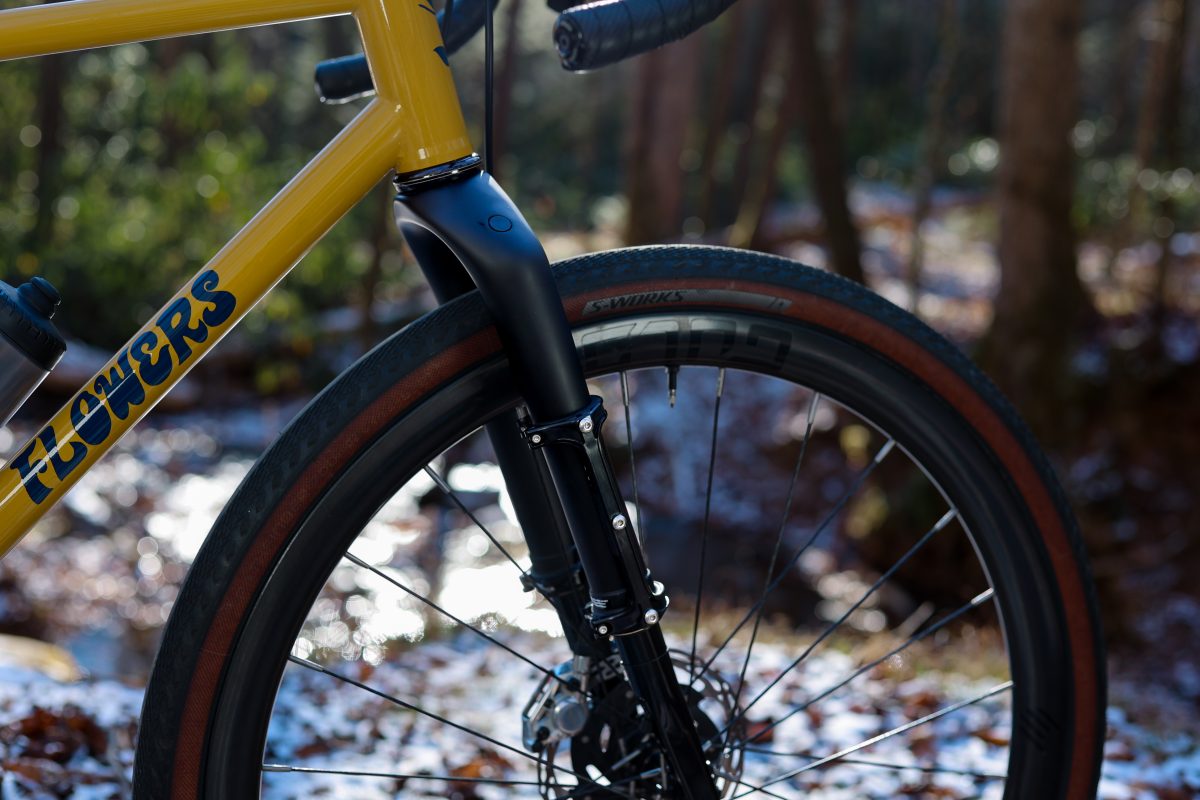 FLOWERSと書かれた黄色い自転車の前輪とフォークのクローズアップ。背景は浅い森で、地面には雪が積もっている。.