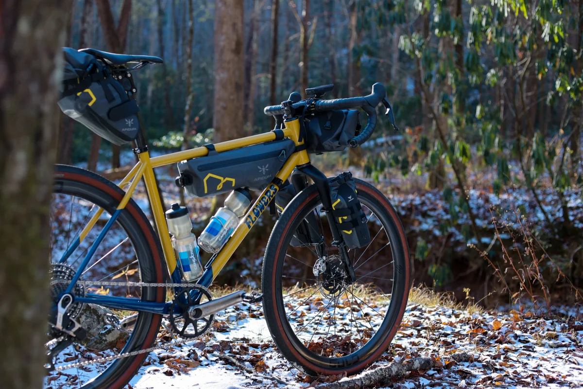 木々と落ち葉に囲まれた雪の林道に、複数のバッグと水筒を積んだ黄色いツーリング自転車が立っている。.