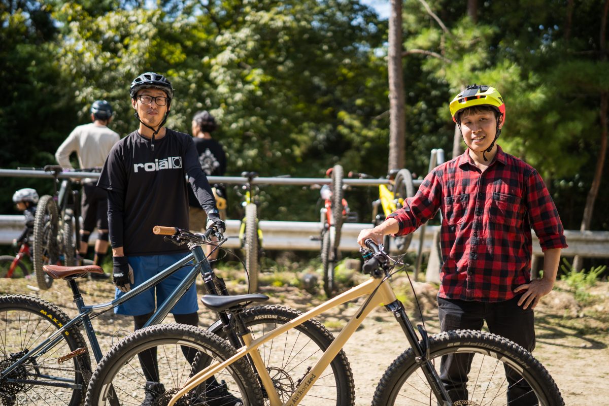 2人の男性がマウンテンバイクを手に、トレイルに立っている。背景には、他のサイクリストや自転車が見える。.