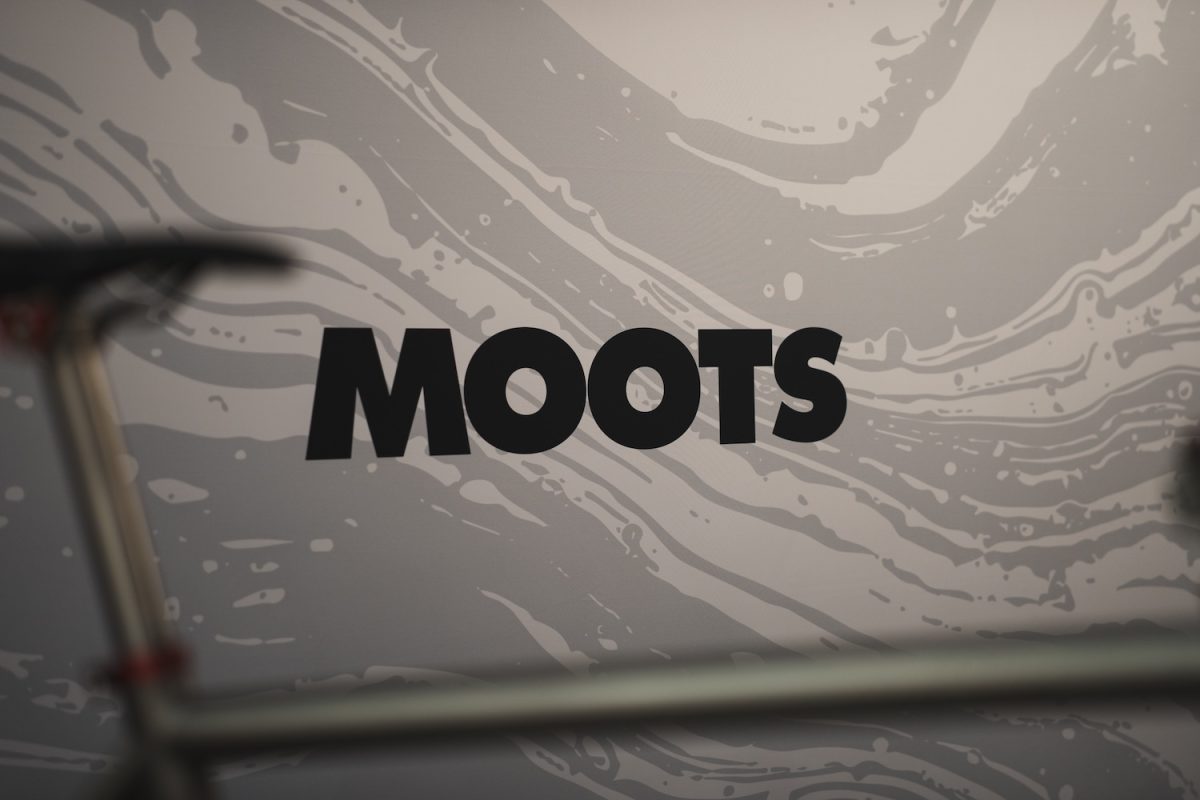 グレーと白の渦巻き模様の背景に、黒い文字でMOOTSと書かれており、その一部が自転車のフレームに隠れている。.