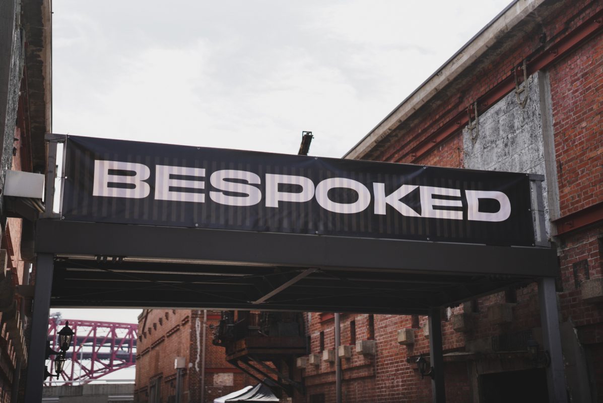 2つの赤レンガ造りの建物の間に、白い文字でBESPOKEDと書かれた黒い横断幕が掲げられている。.