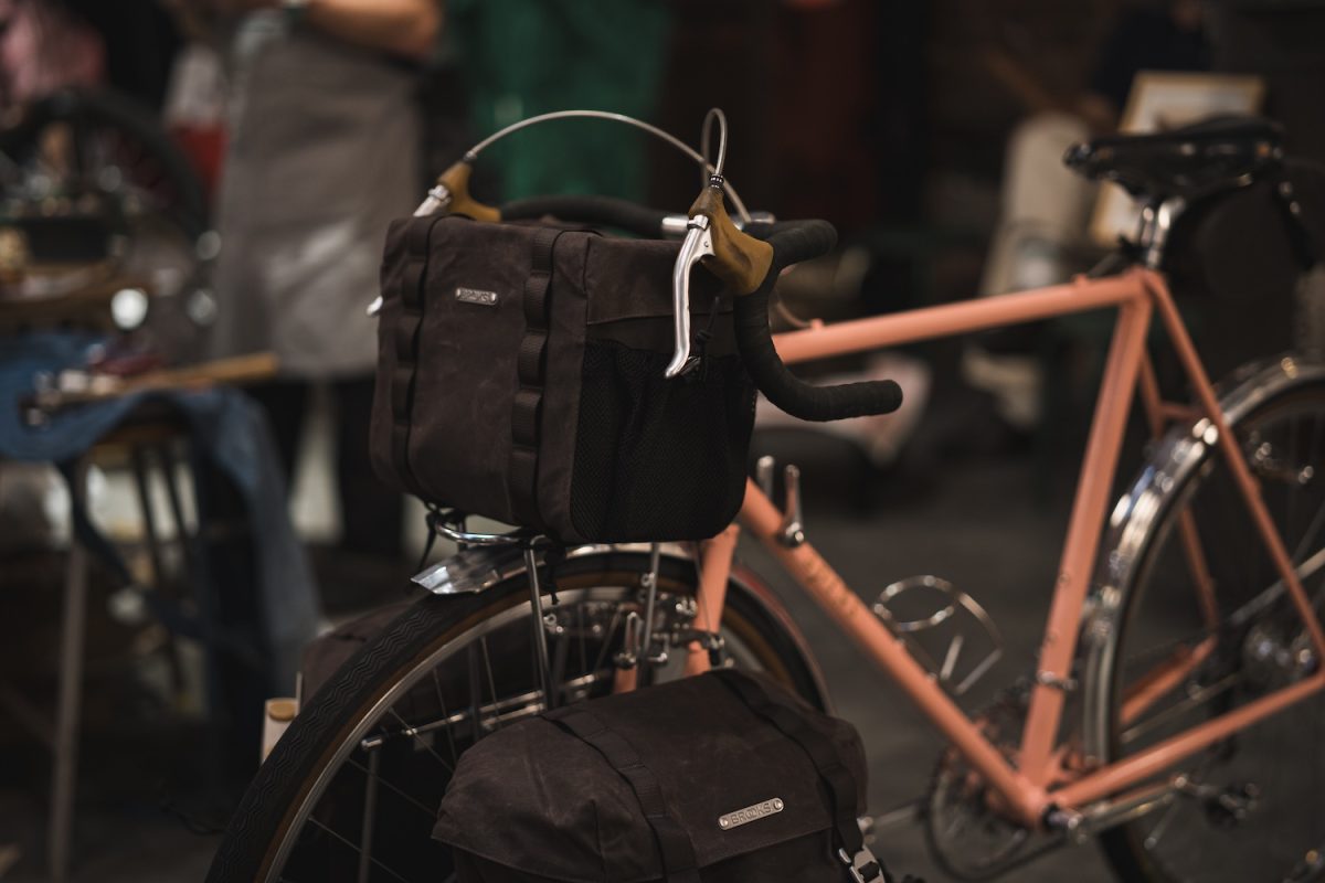 ピーチ色の自転車に、ブルックス製のハンドルバーバッグと、それに合わせたリアラックバッグが取り付けられています。.