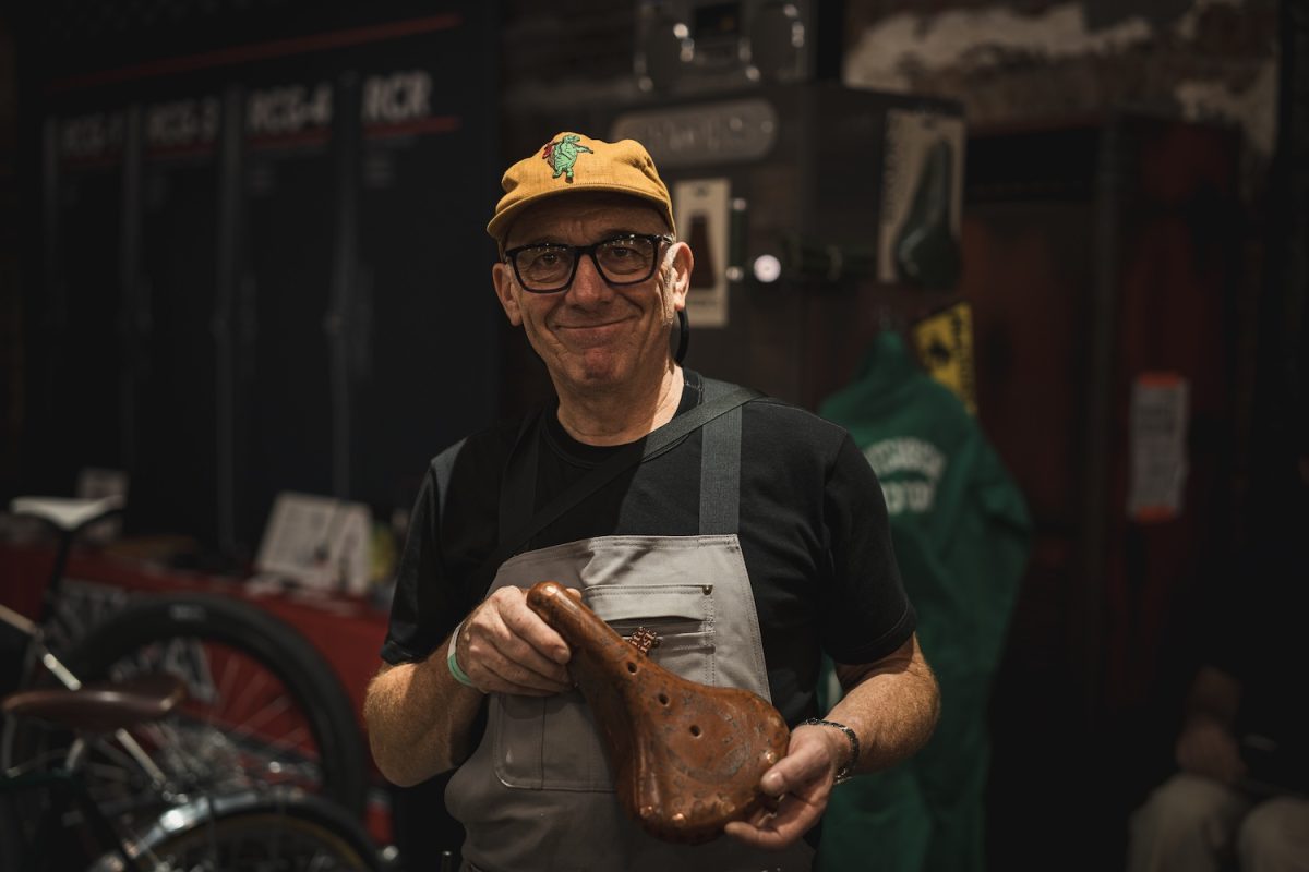 眼鏡をかけ、黄色い野球帽をかぶり、茶色の革製の自転車用サドルを抱えている男性。.