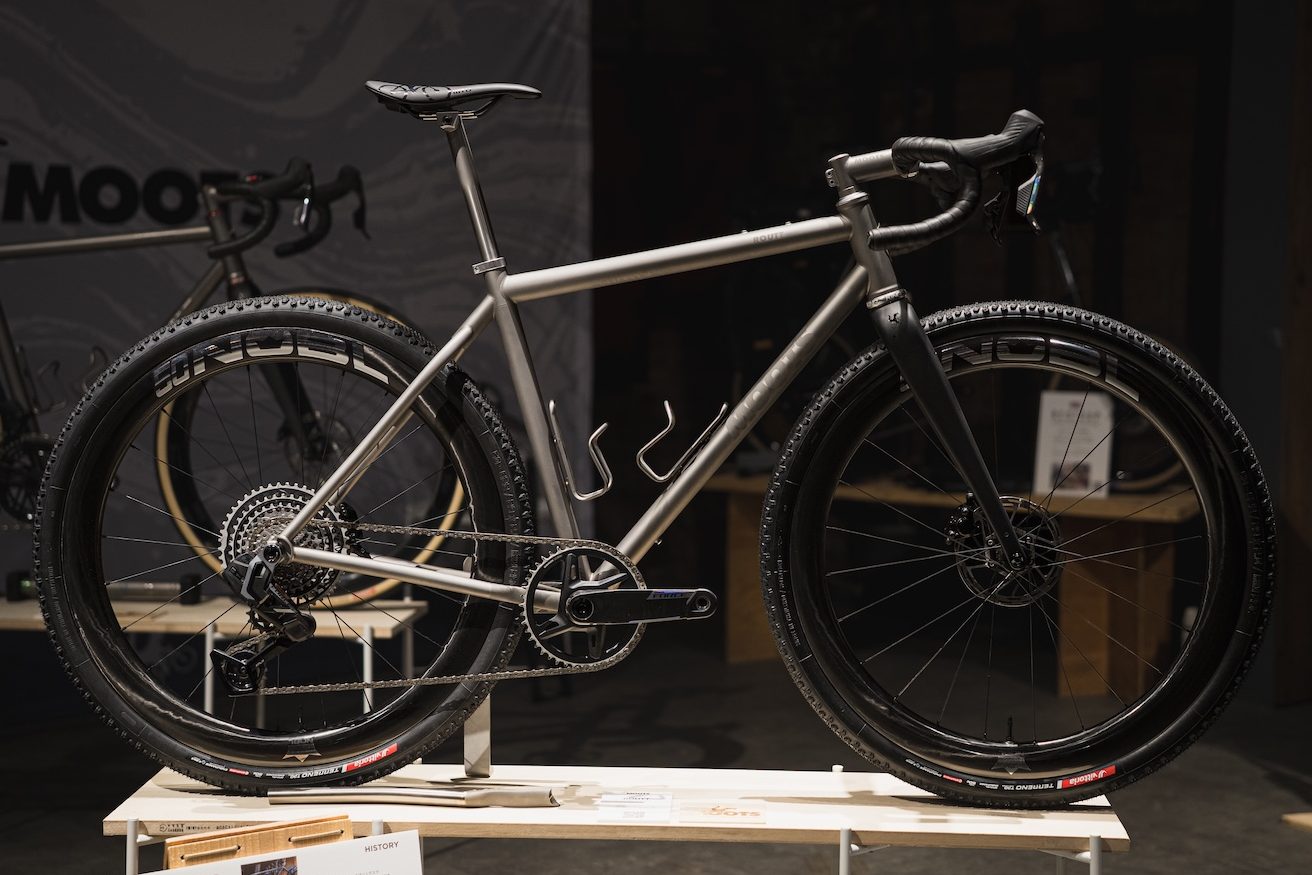 ドロップハンドル、カーボンフォーク、幅広のノビータイヤを装備したMootsのチタン製グラベルバイクが、展示台に置かれている。.