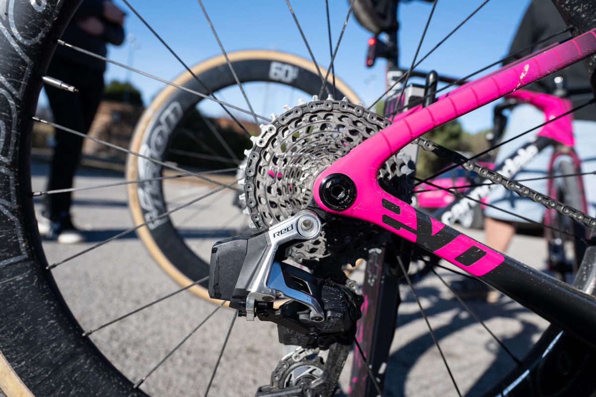 ピンクの自転車のリア・ディレイラーとギアのクローズアップ、背景には別の自転車と人々。.