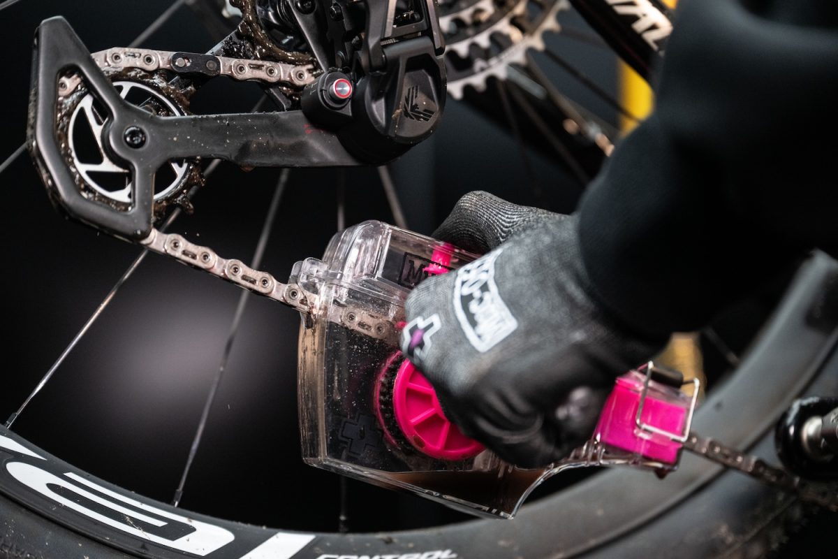 手袋をはめた人が、チェーン清掃用具を使って自転車のチェーンとギアを清掃している。.