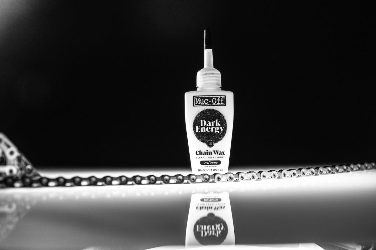 ノズルが尖ったMuc-Off Dark Energy Chain Waxのボトルが、自転車チェーンの横の反射面に直立している。.