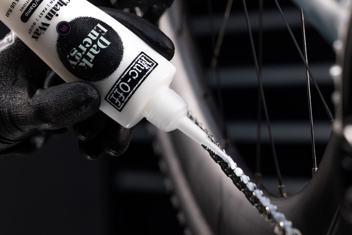 手袋をはめた手で、Dry Lubeと書かれたボトルから白い液体を自転車のチェーンに塗る。.