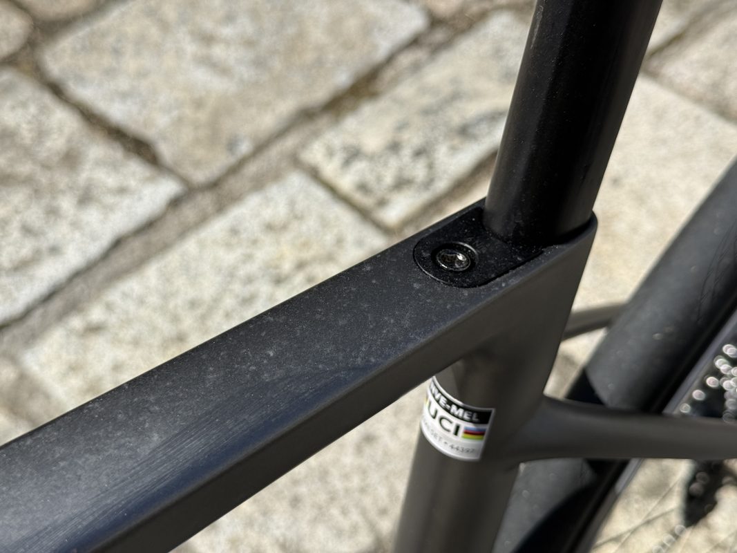 石畳を背景に、マットブラック仕上げ、ファスナー、UCI認証ステッカーを示す自転車フレームのクローズアップ。.