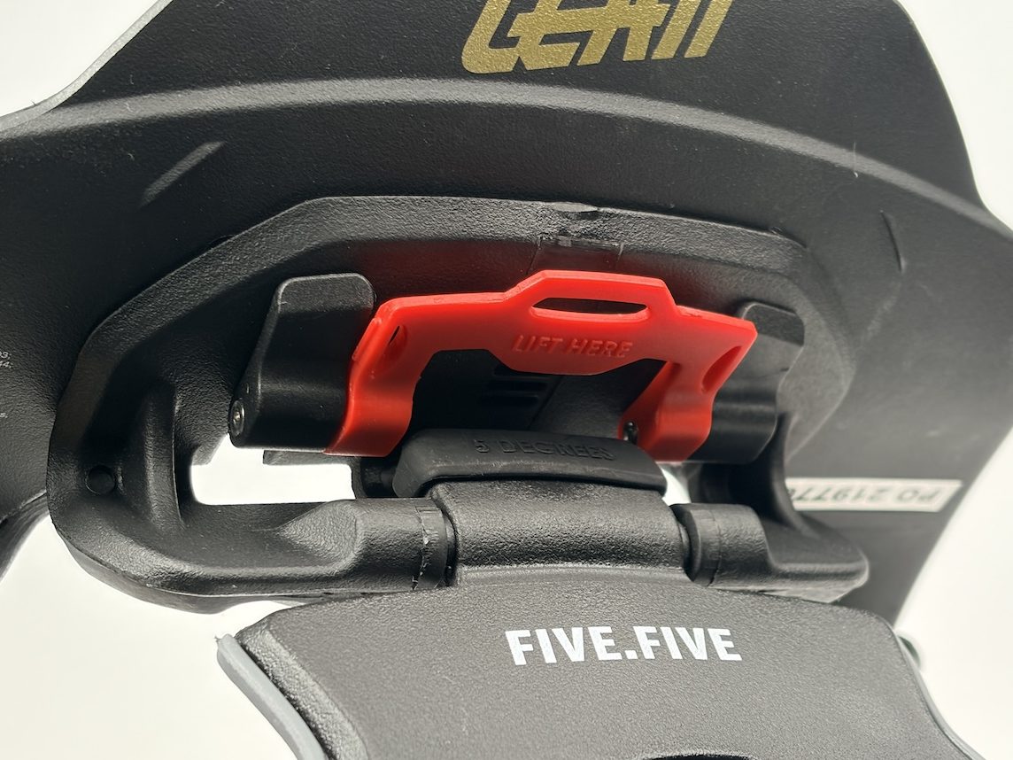 黒いLeatt製ネックブレースのクローズアップ写真。赤いLIFT HEREのラッチ機構とFIVE.FIVEのラベルが確認できる。.