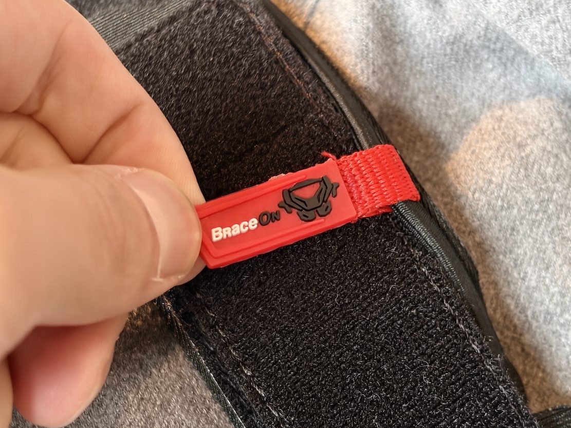 ある手が、マジックテープのストラップに取り付けられたBrace Onブランドの小さな赤いタグを引っ張っている。.