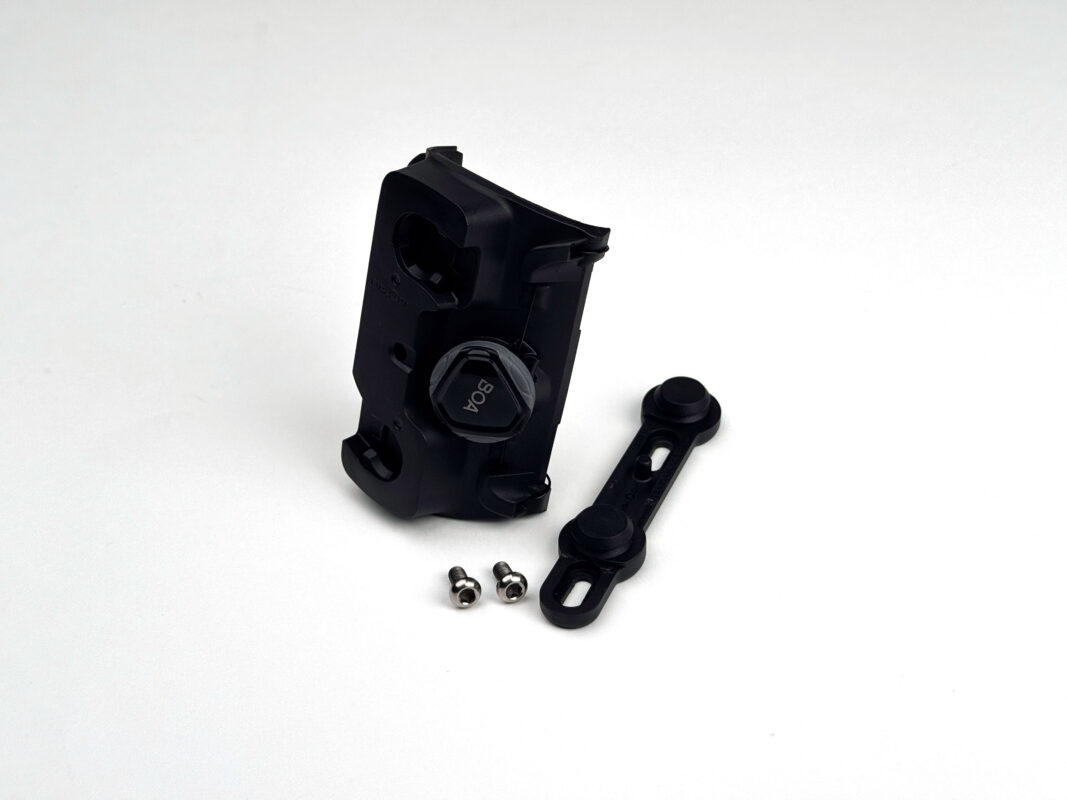 ツイスト・ユニコネクター＋バイクベース（TWIST UNI CONNECTOR + BIKE BASE）は、黒いプラスチック製で、BOAダイヤル、2本の小さなネジ、取り付けストラップが白い表面に表示されている。.