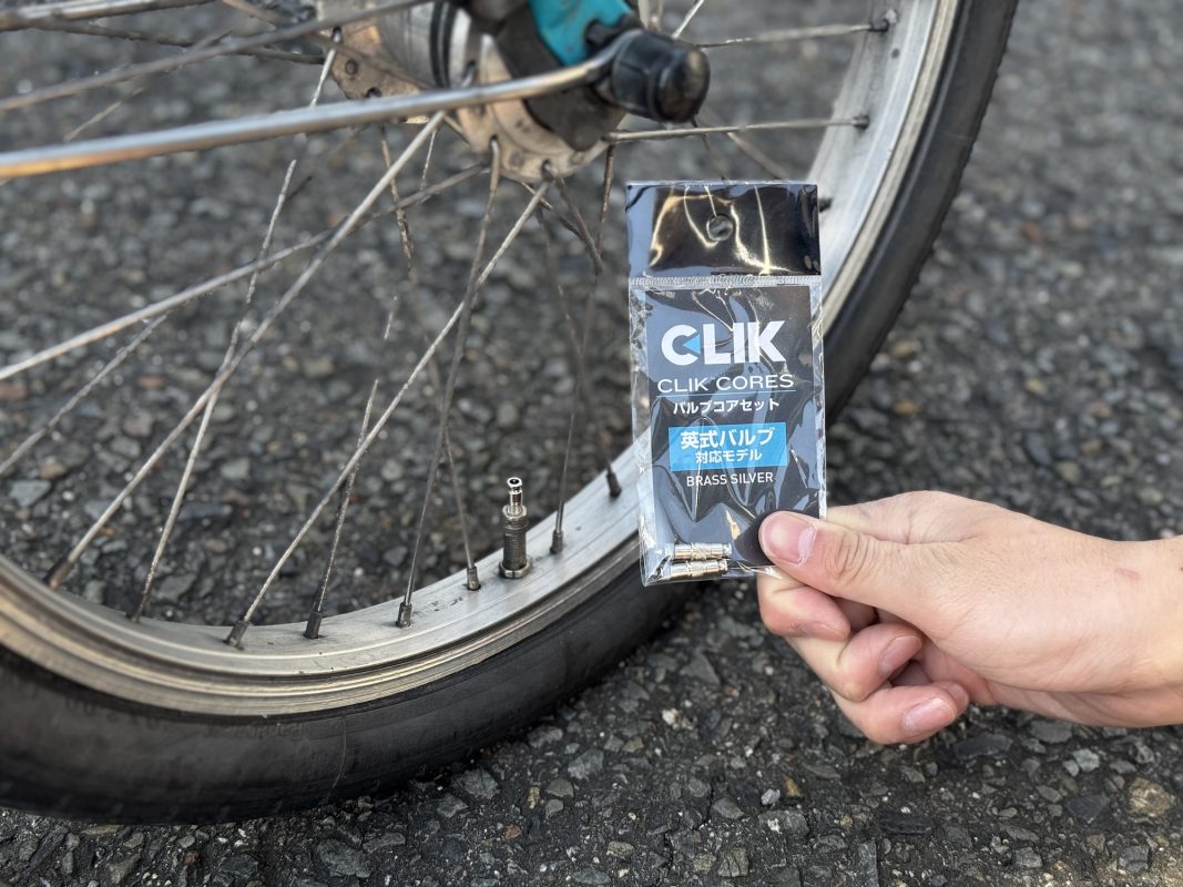 舗装された路面に置かれた自転車の車輪のバルブ付近で、「Clik Cores」と書かれた包みを持つ手。.