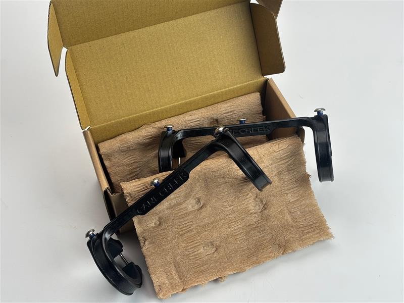 CANE CREEKと書かれた2つの黒い金属製の自転車部品が、開いた段ボール箱の中の褐色のパッドの上に置かれている。.