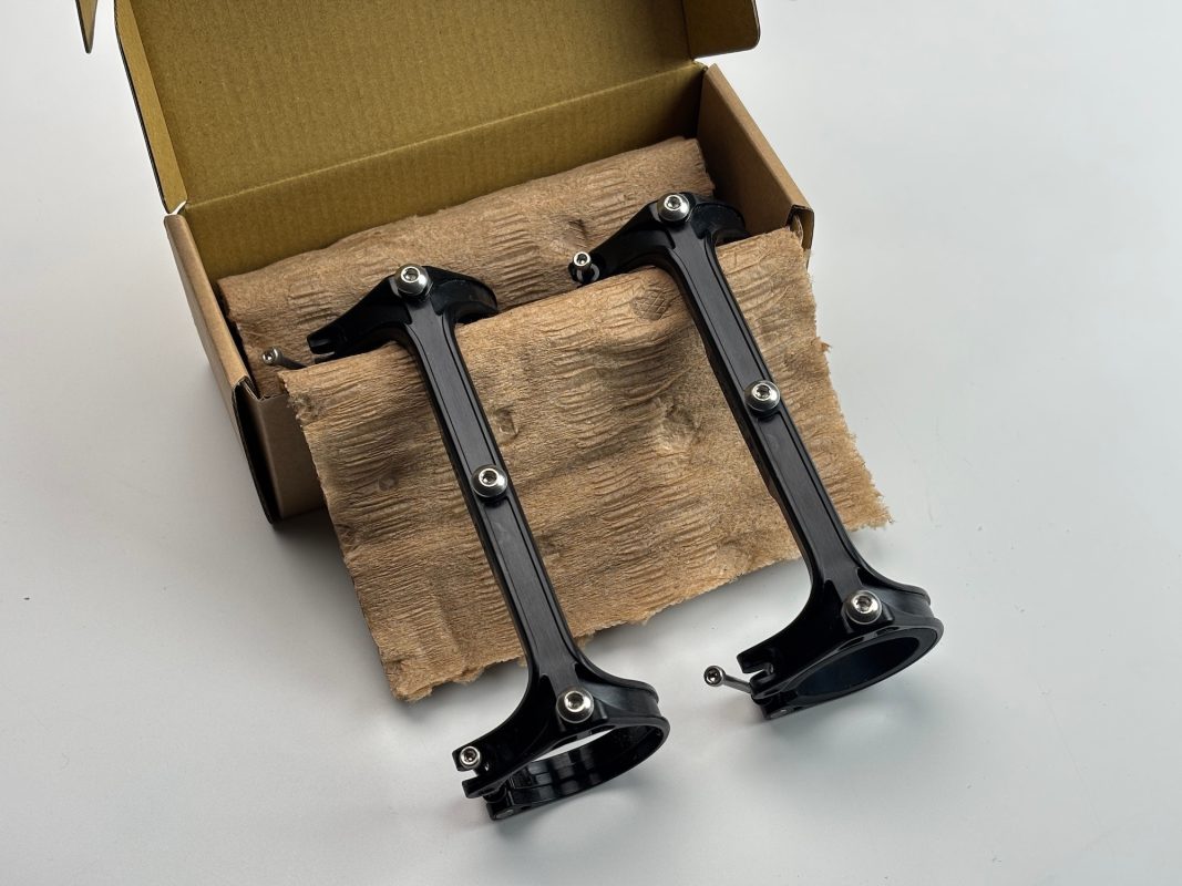 2つの黒い金属製ブラケットとスクリュー・マウントが、白い表面の開いた茶色の箱の中に部分的にある、成形された段ボールのパッケージの上に置かれている。.