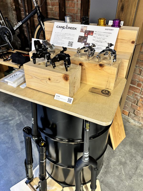 黒い樽の上に置かれた木製の台座に、ケーン・クリークの自転車用ブレーキ部品が展示されており、その横には同社の沿革を説明する案内板が掲げられている。.