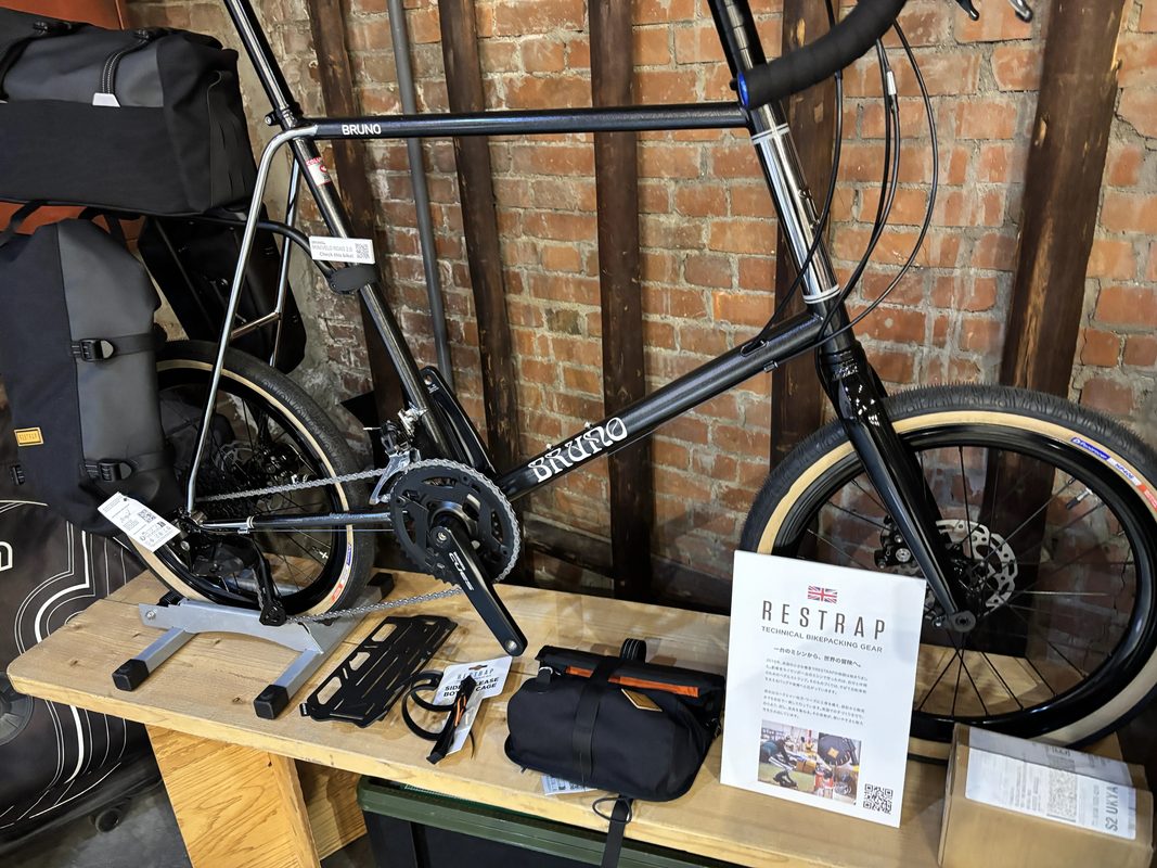 レンガの壁を背景に、ディスプレイスタンドに設置された黒いブルーノの自転車。その前の木製の棚には、Restrap製のバイクパッキングバッグと説明看板が置かれている。.