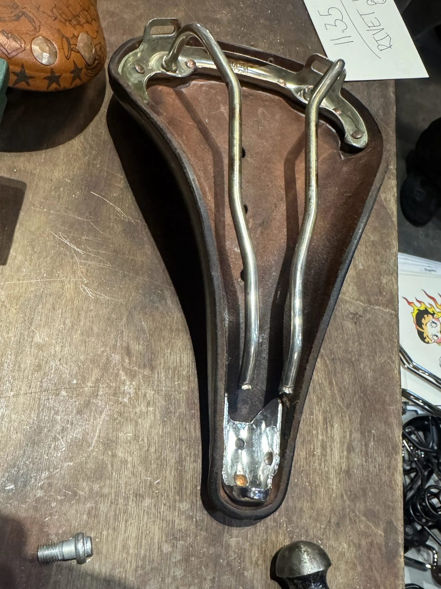 作業台の上に、金属製のレールが付いた革製の自転車用サドルが置かれており、その隣にはハンマーと、11:35 RIVET MANと書かれたメモがある。.