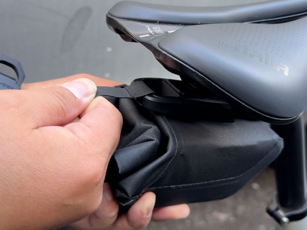 ある人がストラップを引っ張り、自転車のサドルの下に黒いサドルバッグを固定している。.