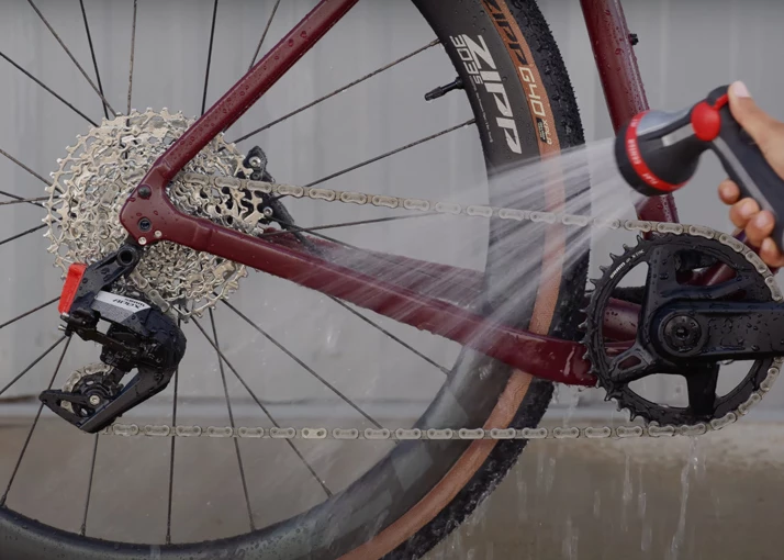 ある人がスプレーノズルを使って、マルーン色の自転車のリアディレイラー、カセット、チェーンを洗い流している。.