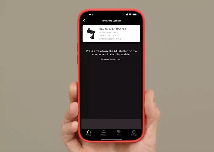 ある手が、RED RD XPLR MAX 46Tコンポーネントのファームウェア更新画面が表示されたスマートフォンを持ち、ユーザーにAXSボタンを押して開始するよう指示しています。.