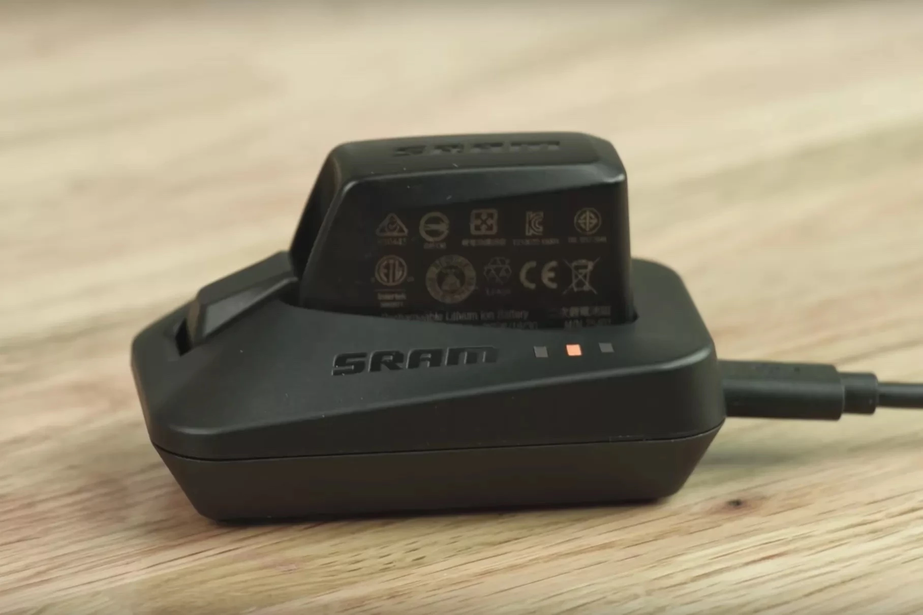 バッテリーが挿入された黒いSRAM製バッテリー充電器で、オレンジ色のインジケーターランプが点灯しており、側面にUSBケーブルが接続されている様子。.