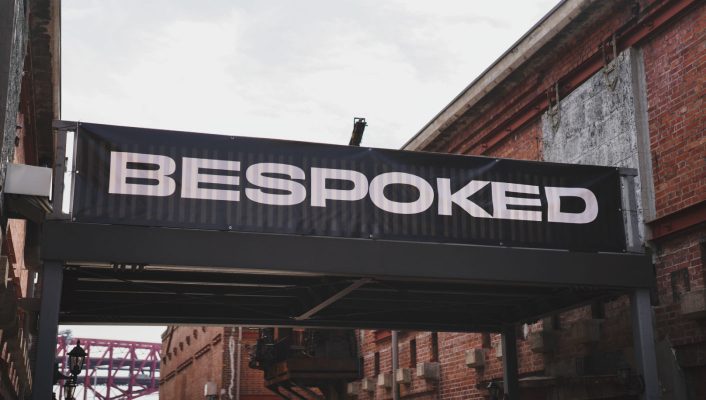 2つのレンガ造りの建物の間に、白い大文字でBESPOKEDと書かれた黒い横断幕が掲げられている。.