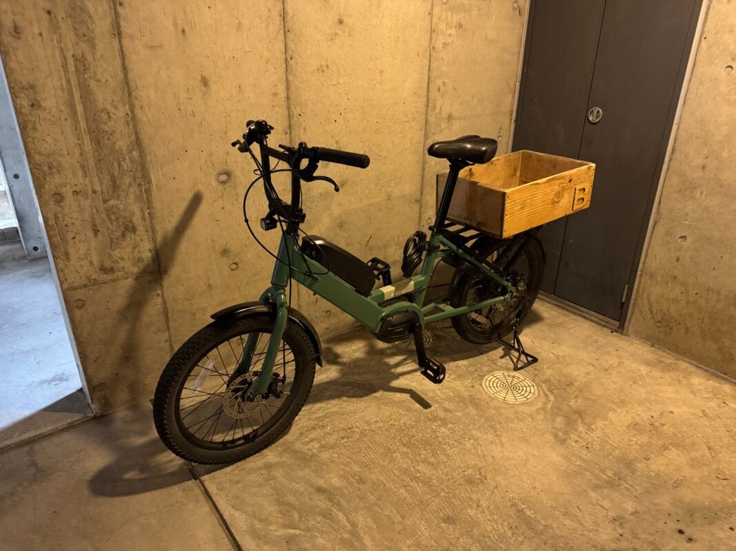 後部ラックに木製の箱が載せられた緑色の電動自転車が、コンクリートの壁に寄りかかって停められている。.