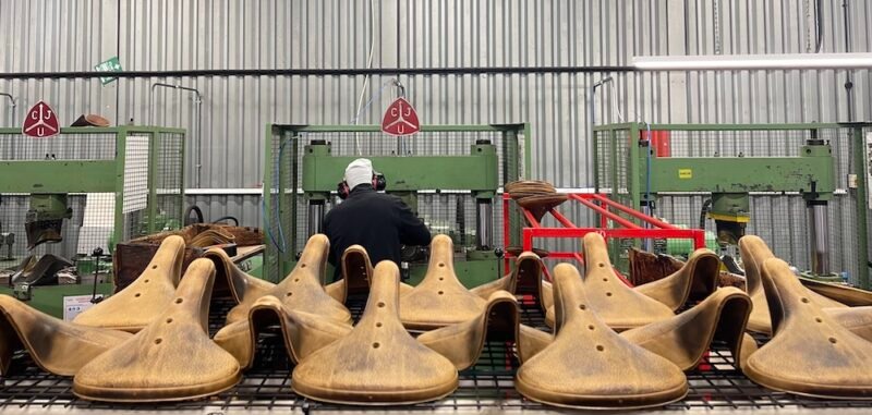 工場内のラックに、茶色の自転車のサドルがずらりと並んでおり、その背景では作業員が機械を操作している。.