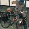 imgi_37_c514b146ebd92abefaba3d34b2420a9045a61f6b_Travelling_Bags_Rambler_Isle_of_Wight_Safe_sound_and_easily_accessed 帽子とショートパンツ姿の男性が、茶色のキャンバス製パニアバッグとRAMBLER RAND BAG SEAL BROWNのハンドルバーバッグを装備した、荷物を満載したツーリングバイクにもたれかかっている。.