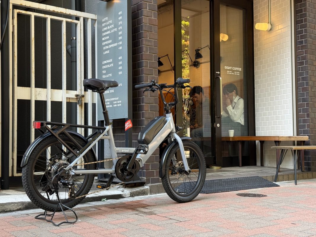 メニューボードが掲げられたコーヒーショップ前の歩道に、銀色の電動自転車が停まっている。.