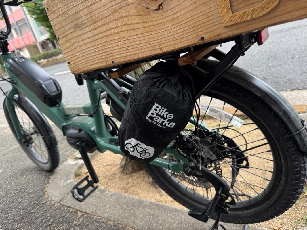 緑色の電動自転車のリアラックに、黒いバイクパーカー型の自転車カバーバッグがぶら下がっている。.