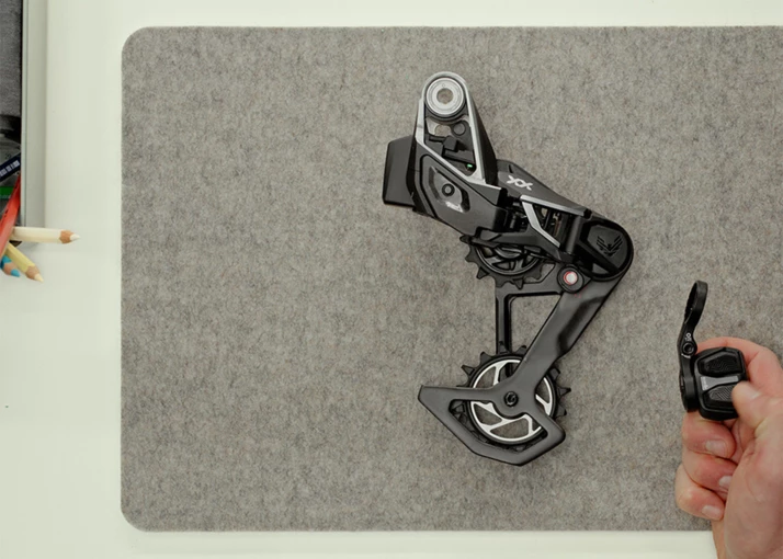 灰色のフェルトマットの上に置かれたSRAM XXのマウンテンバイク用リアディレイラーが、手に持たれている。.