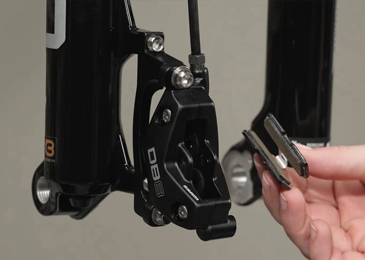 自転車のフォークに取り付けられたSRAM DB8ディスクブレーキキャリパーのそばで、自転車のブレーキパッドを2枚持った手。.