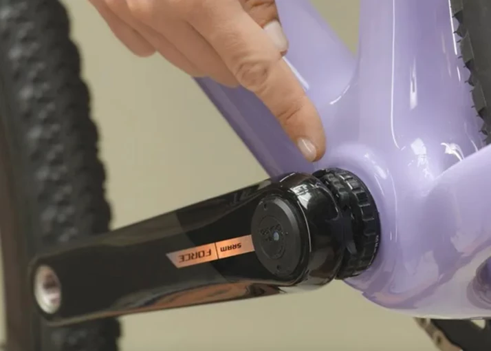 黒いSRAM Forceクランクセットが取り付けられた、ラベンダー色の自転車フレームのボトムブラケット部分を指さしている指。.