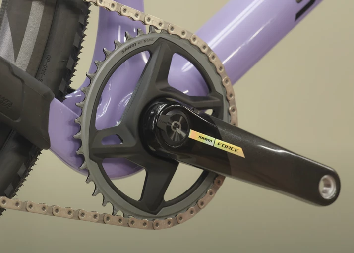 淡い紫色のフレームに、SRAM Forceのクランクセット、シングルチェーンリング、そして金属製のチェーンを備えた自転車のドライブトレインのクローズアップ。.